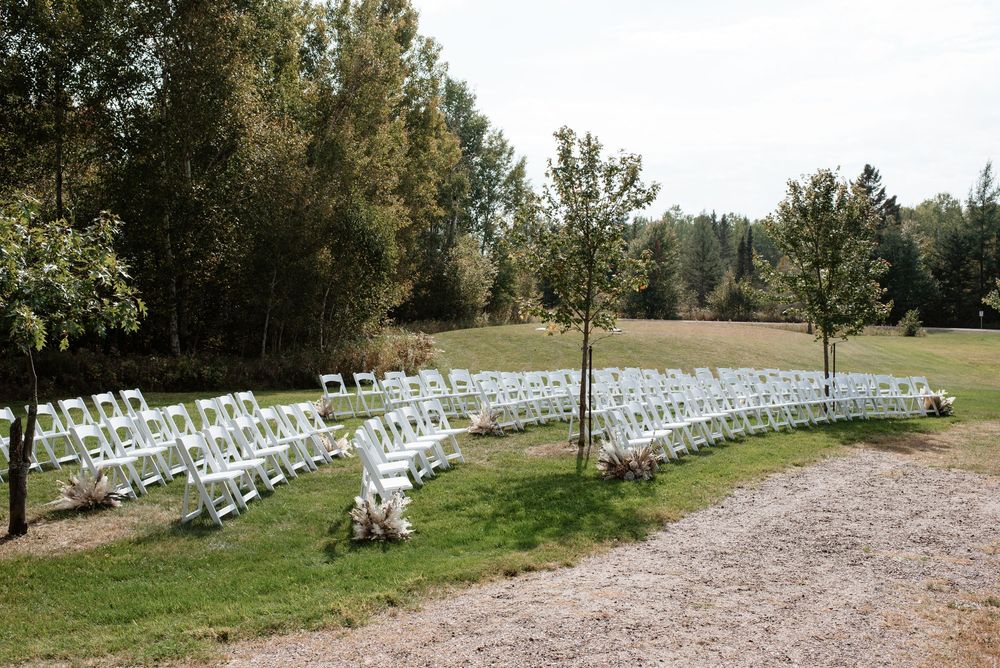 White folding chairs arranged in curved rows for an outdoor wedding ceremony surrounded by lush green trees and grass.