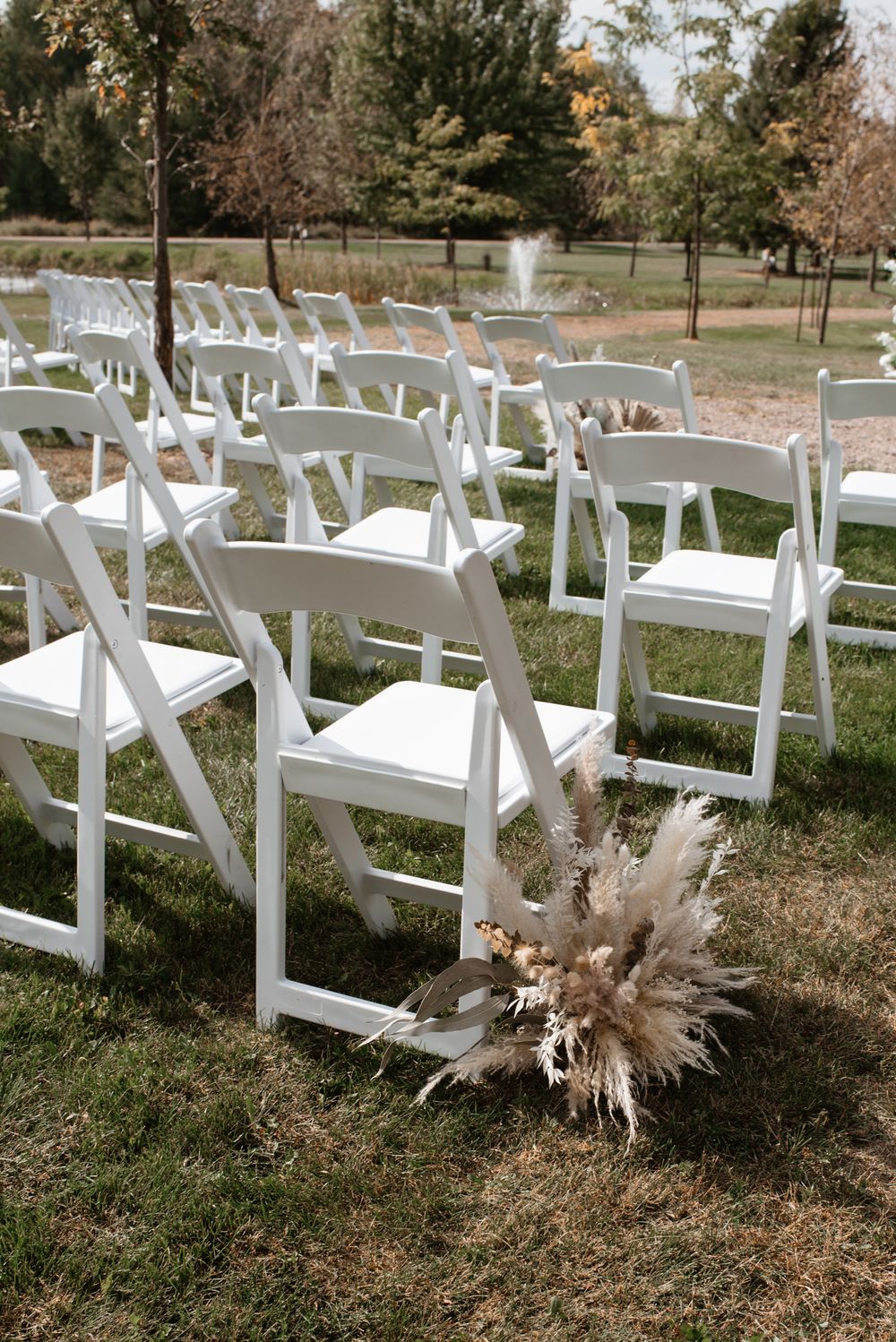White folding chairs arranged in rows on grass for an outdoor wedding ceremony with pampas grass bouquet aisle decor.