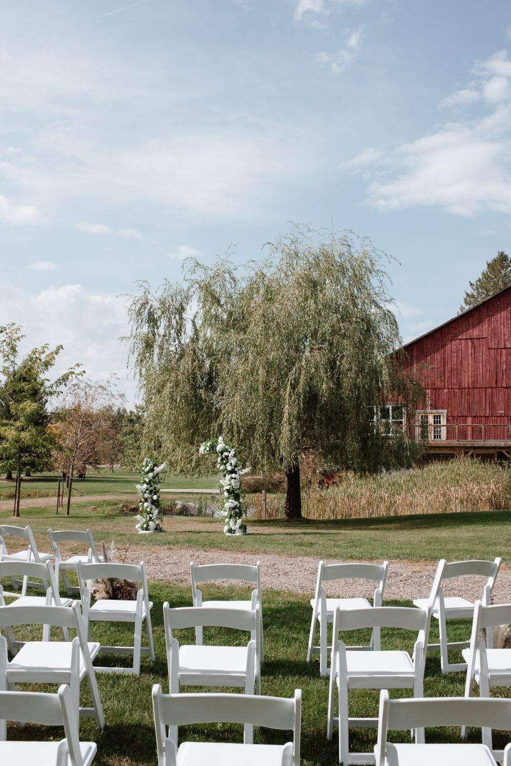 Outdoor wedding ceremony setup with white folding chairs, floral arch, weeping willow tree, and red barn on a sunny day.