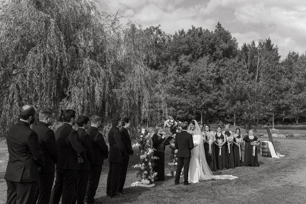 Black and white wedding ceremony outdoors with bridal party under willow trees on a lush green lawn.