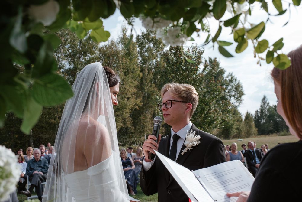 Outdoor wedding ceremony with bride in veil and groom in suit exchanging vows with officiant holding microphone.