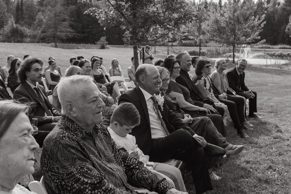 Black and white photo of guests seated outdoors at a garden wedding ceremony, smiling and enjoying the celebration.