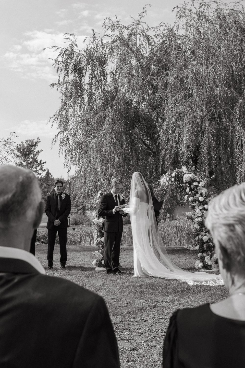Black and white outdoor wedding ceremony under a weeping willow tree with bride in veil and floral arch.