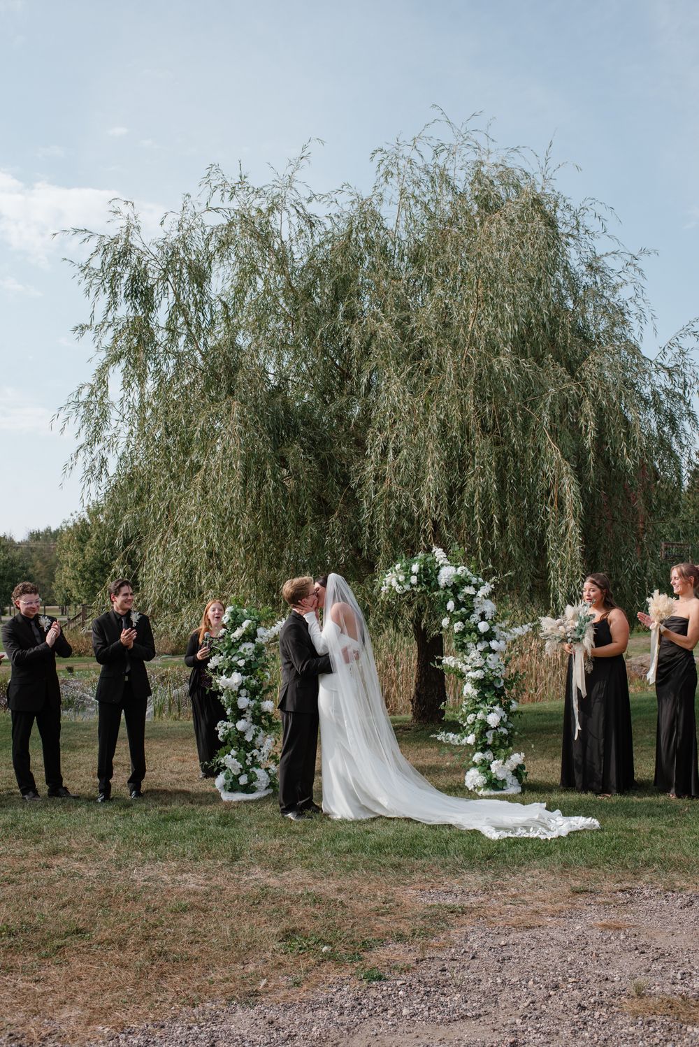 Bride and groom share first kiss at outdoor wedding ceremony under willow tree with floral arch and wedding party in black.