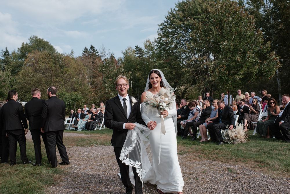 Bride in white gown walks down outdoor wedding aisle with escort on sunny day surrounded by seated guests.