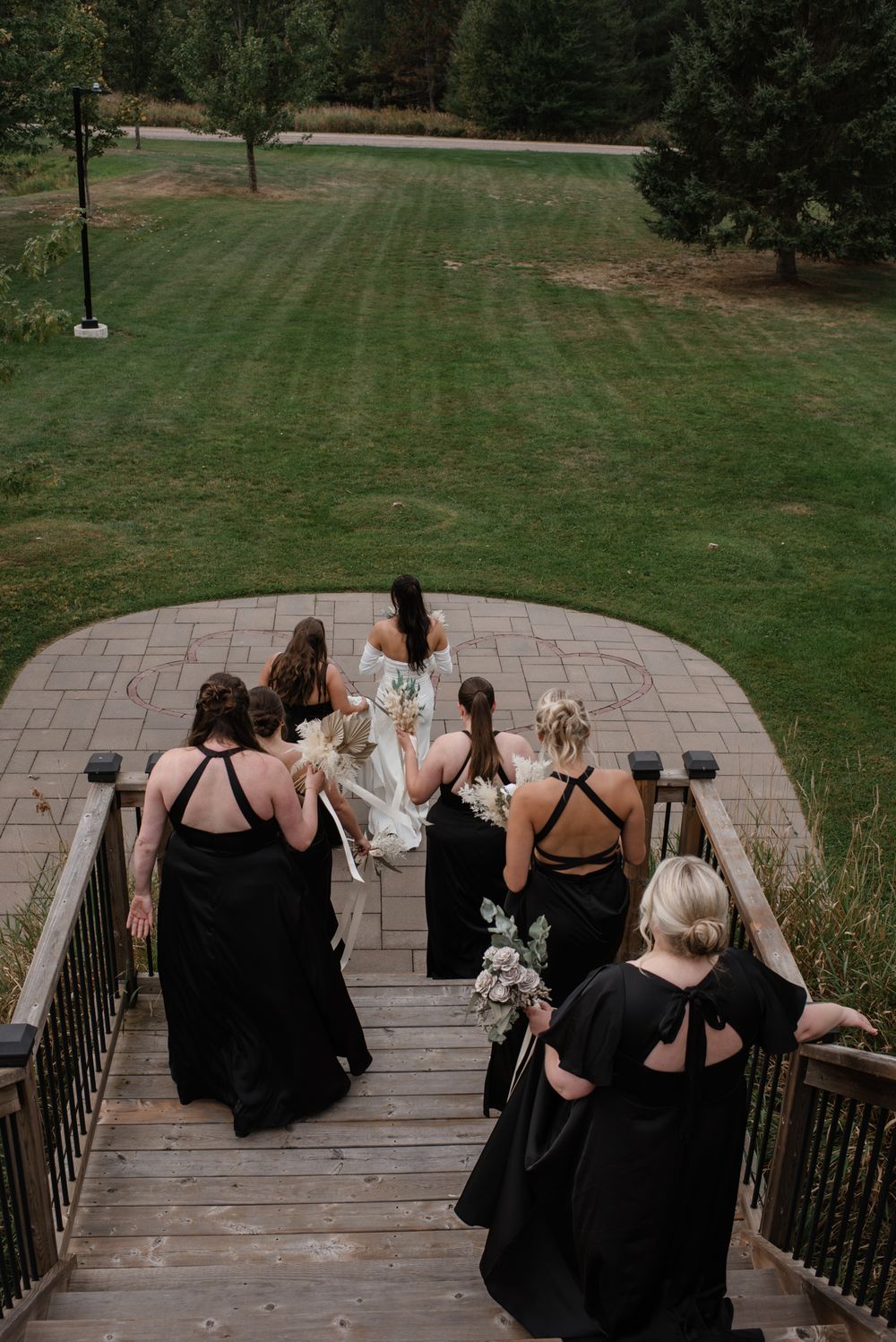 Bride and bridesmaids in black dresses descending wooden stairs toward green lawn at outdoor wedding venue.