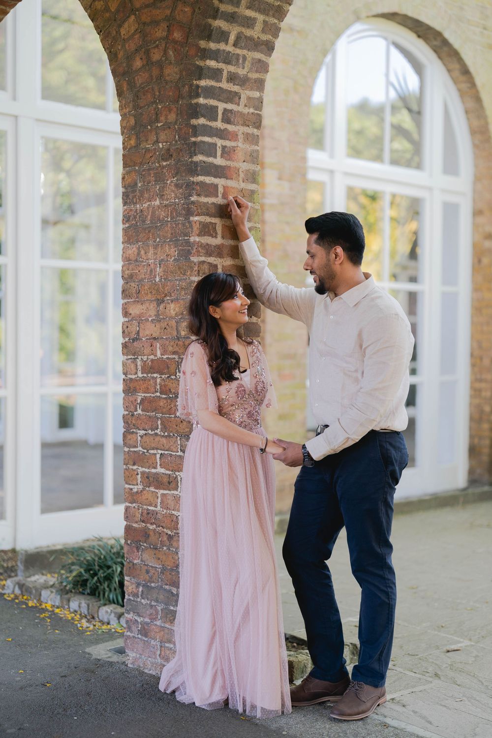 Couple posing romantically by brick archway, woman in pink gown, man in white shirt and navy trousers.