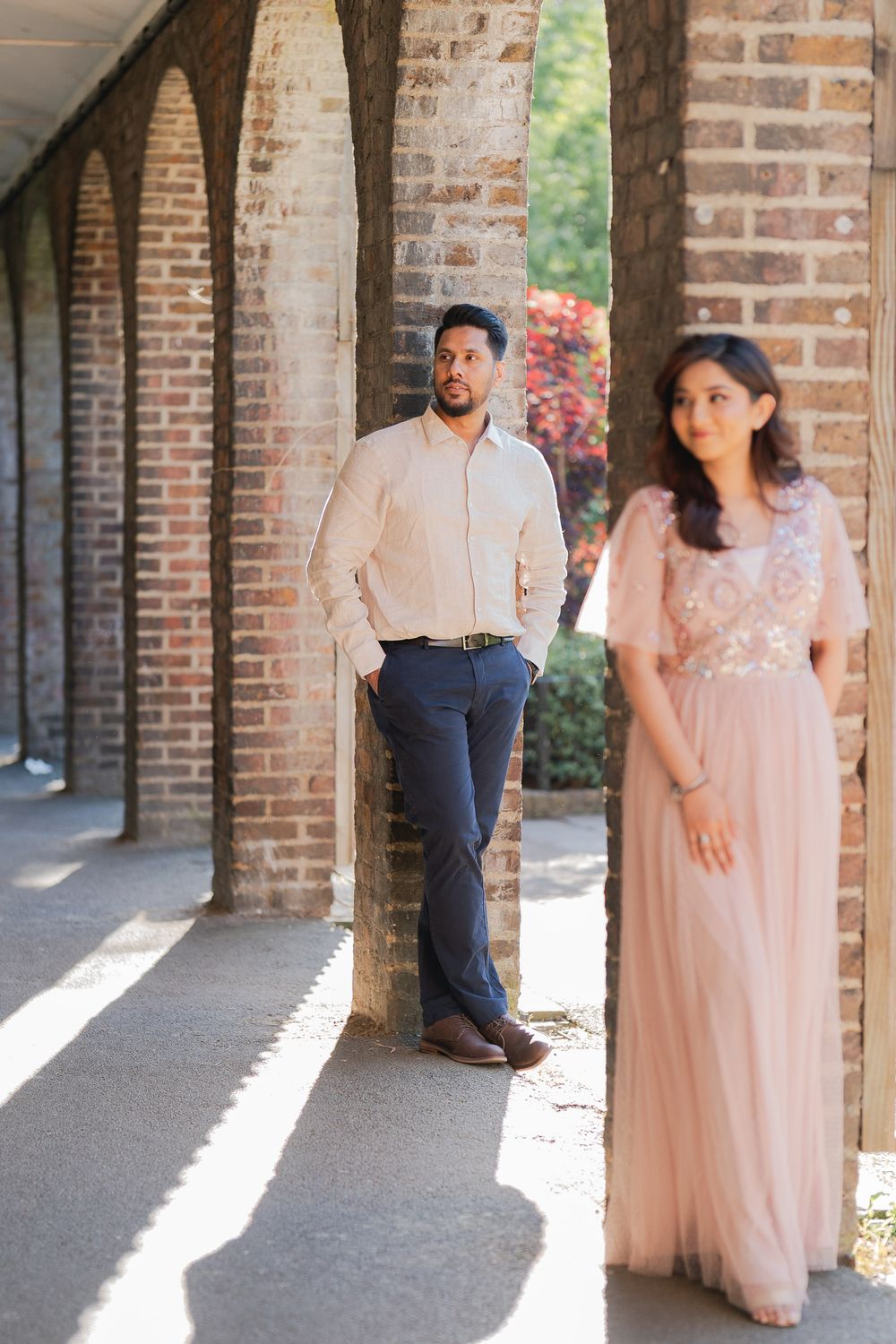 A man in a beige shirt leans against a brick column while a woman in a pink gown stands in the foreground.