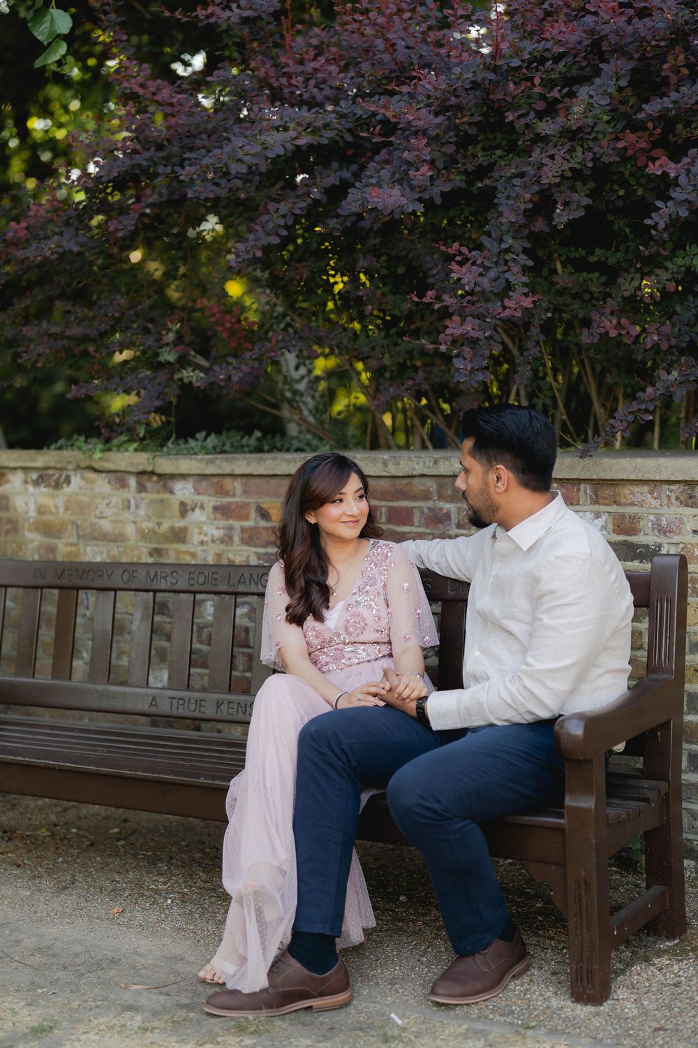 Couple sitting on a wooden bench in a garden, woman in pink dress smiling at man in white shirt.