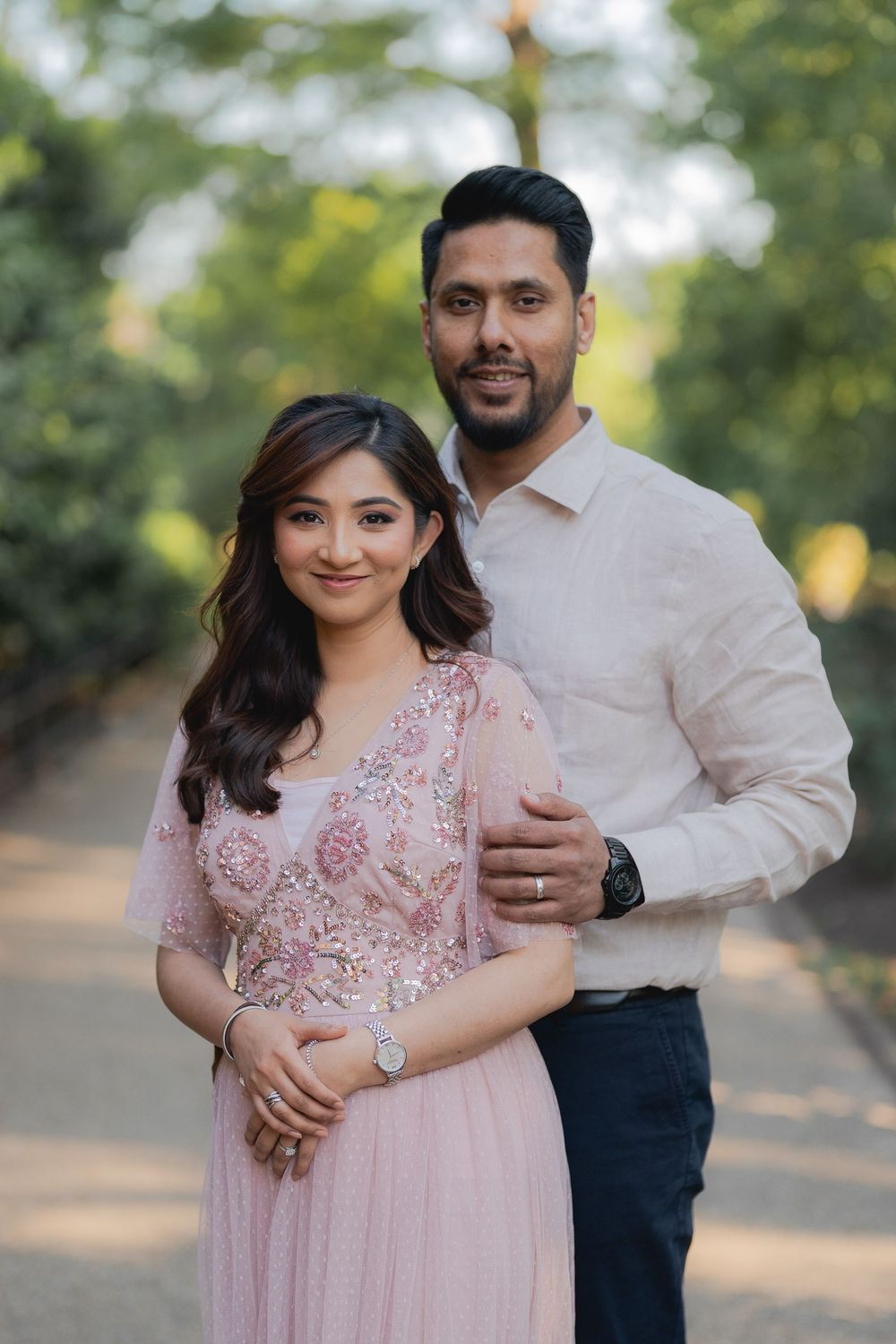 A couple poses outdoors, woman in pink embroidered dress, man in white shirt, surrounded by lush green trees.