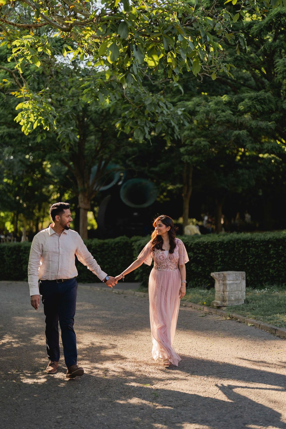 Couple holding hands walking in a lush green park, woman in pink dress, man in white shirt and navy pants.