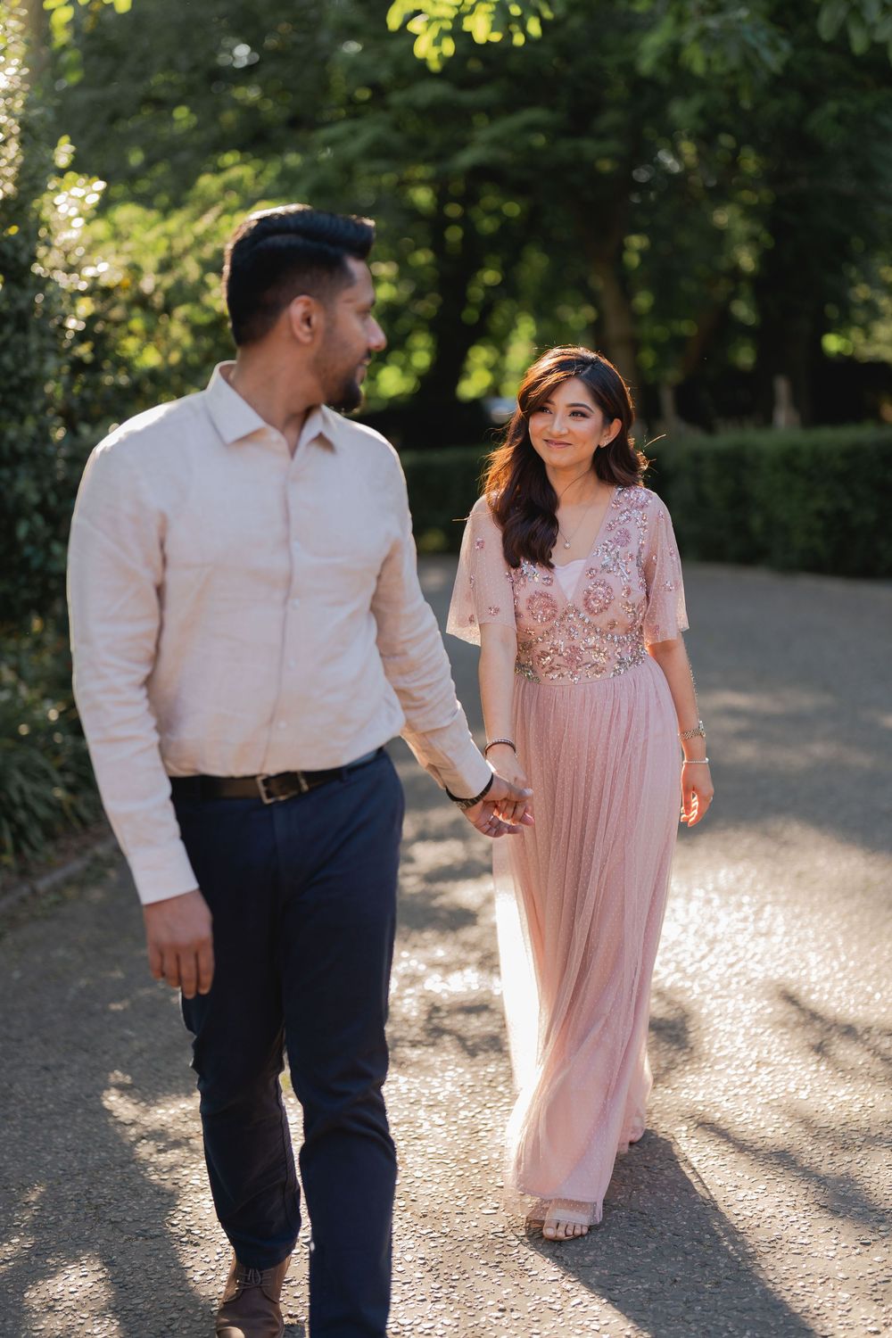Couple walking hand in hand through a sunlit park, woman in pink floral gown smiling at man in white shirt.