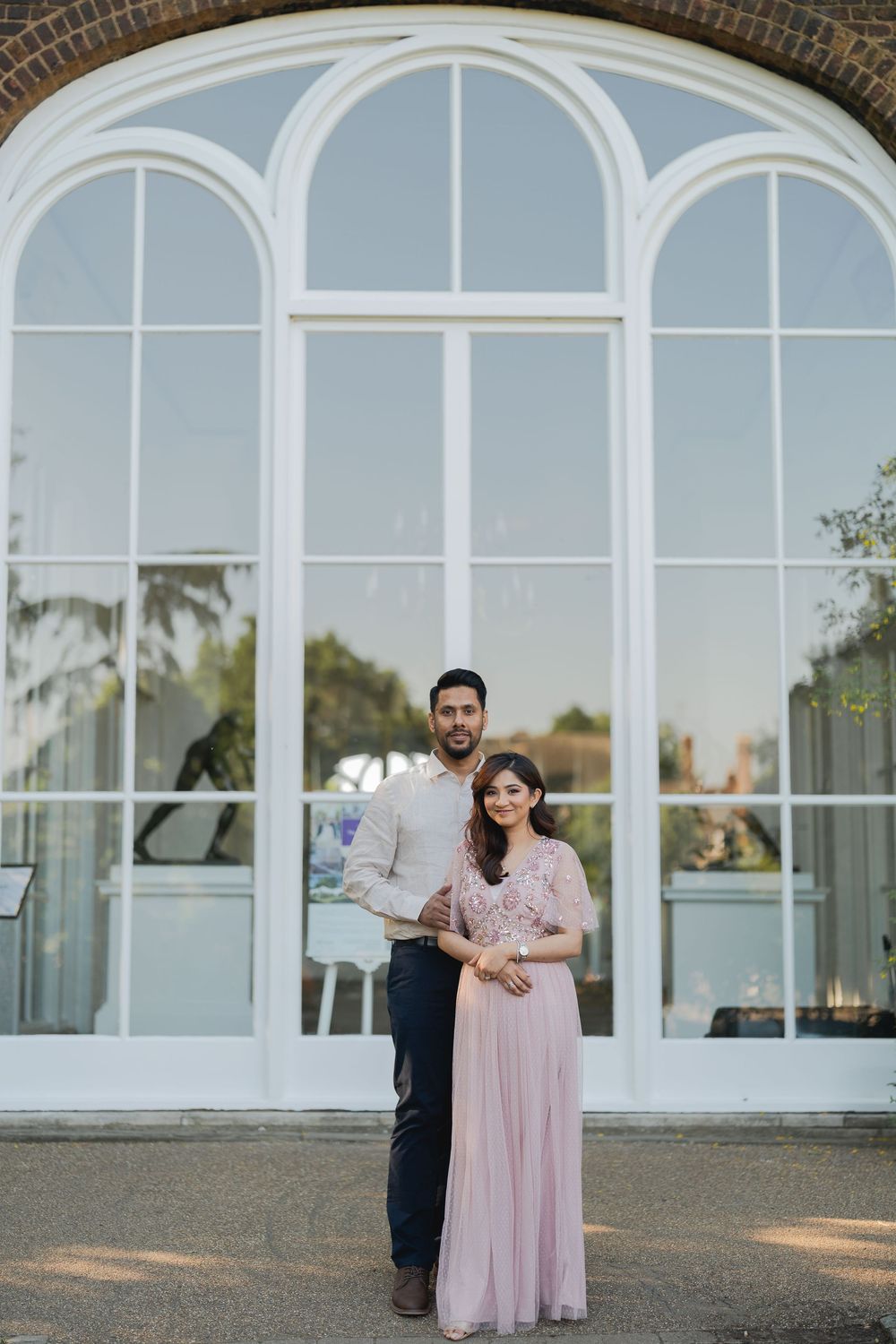 Couple posing in front of elegant arched windows at an outdoor venue, woman in pink gown, man in smart casual attire.