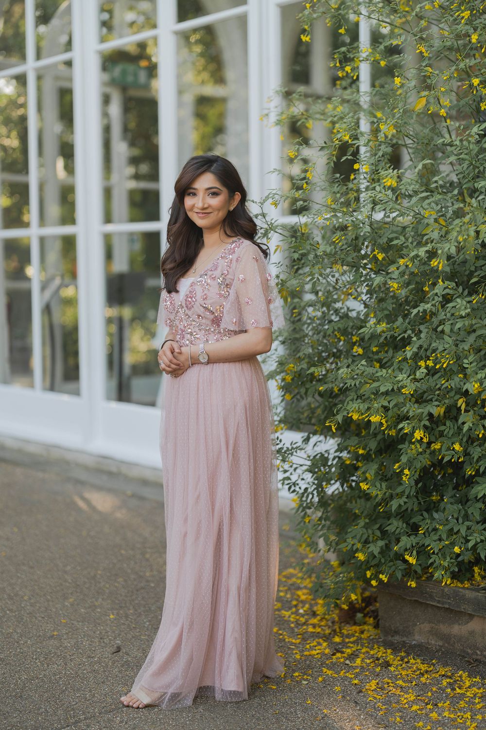 Woman in blush pink embellished maxi dress with flutter sleeves posing outdoors near yellow flowering shrubs.