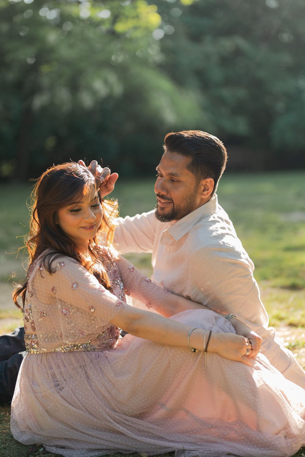 Couple in elegant attire sitting together in a sunlit park, man gently touching woman's hair.