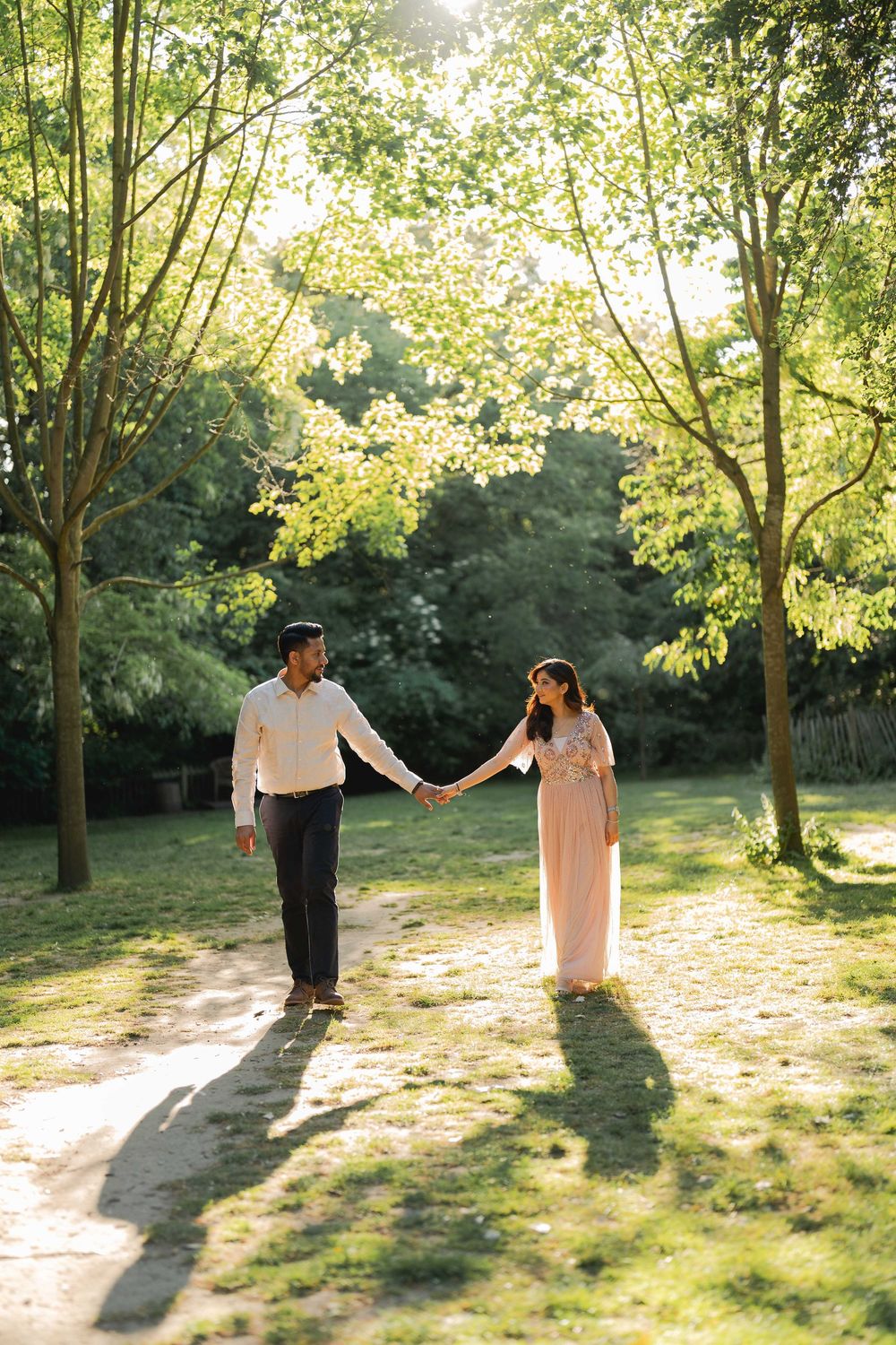 Couple holding hands walking through a sunlit park with lush green trees casting warm golden shadows.