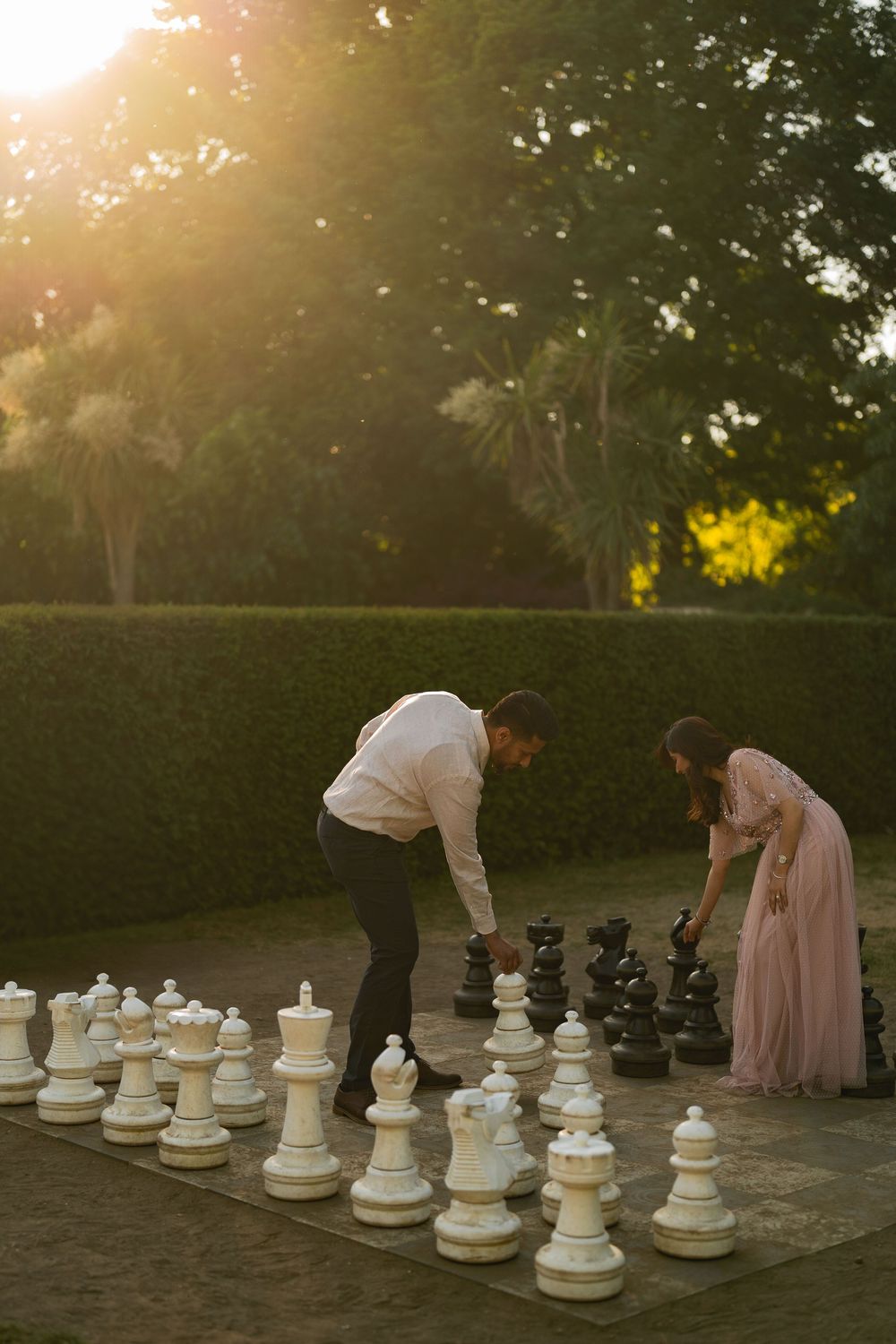 Couple playing giant outdoor chess on stone patio at golden hour sunset with lush green hedges in background.