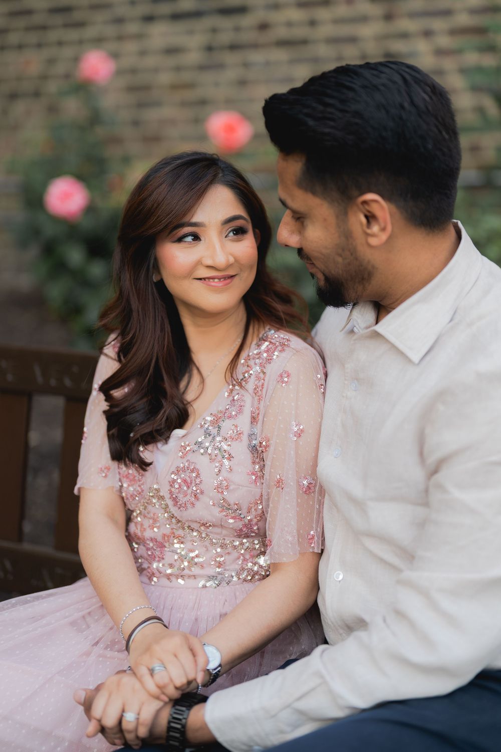 A couple in South Asian attire sits together outdoors, smiling warmly, with pink roses blooming in the background.