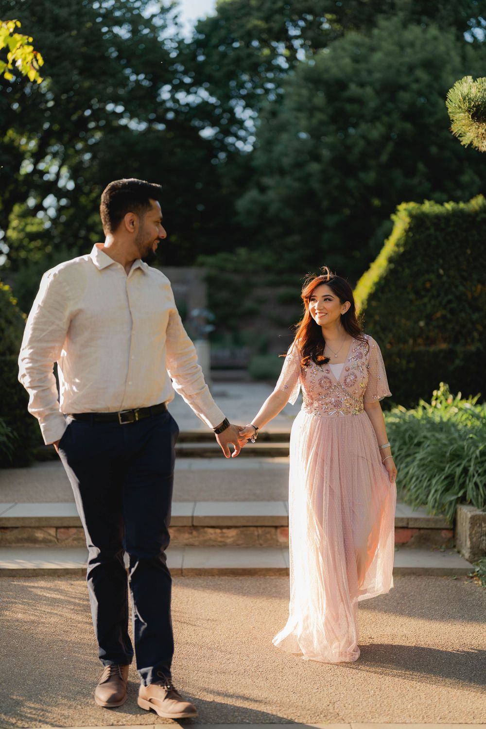 Couple holding hands walking in a garden, woman in pink embroidered gown, man in white shirt and navy pants.