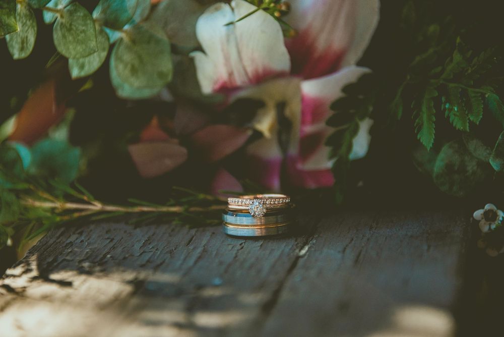 Stacked wedding rings resting on rustic wood surface with colorful floral bouquet backdrop.