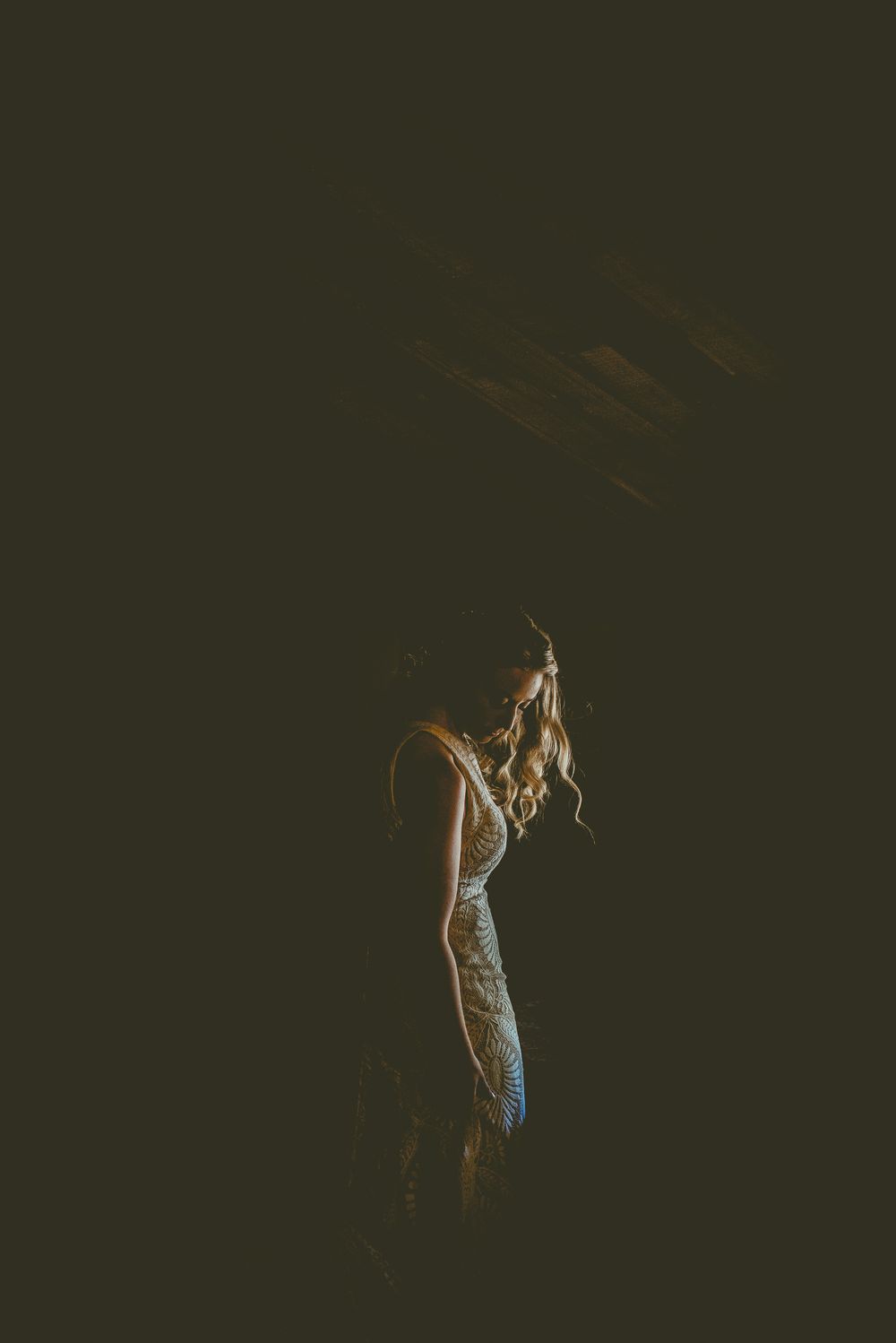 A woman in a white lace micro weddingdress stands in dramatic shadow, illuminated by soft golden light in a dark setting.