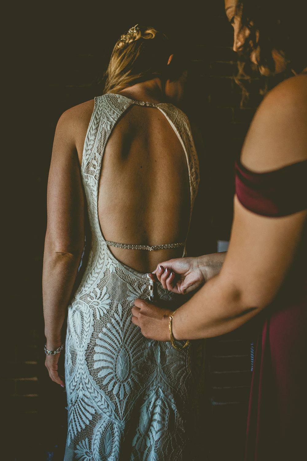 Bridesmaid helping bride fasten open-back lace micro wedding gown with intricate embroidered detail.