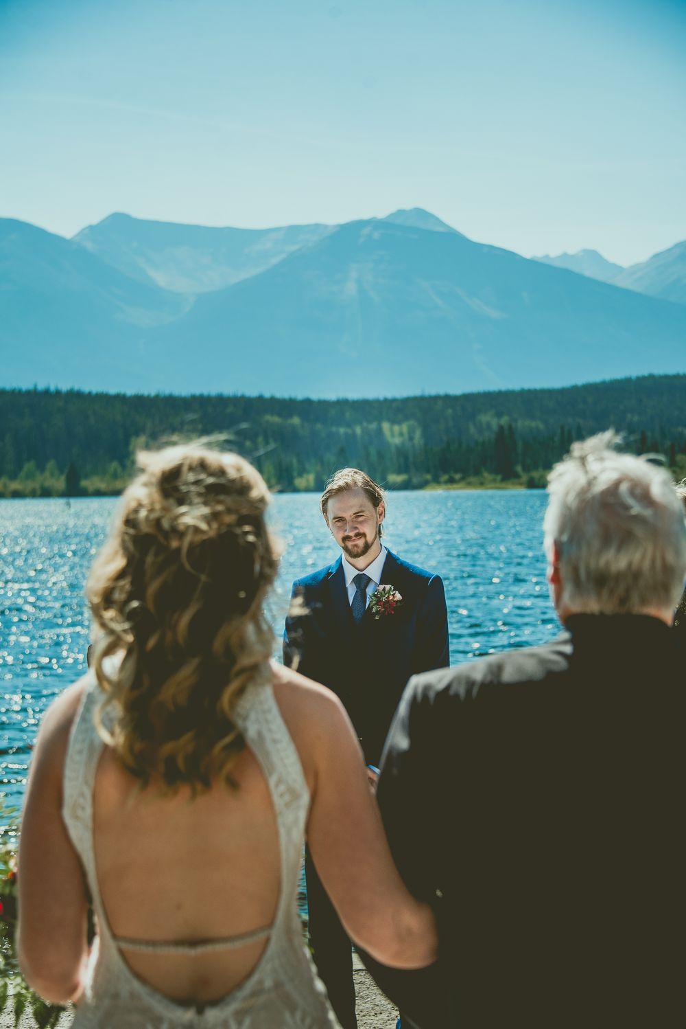 Groom smiling at bride during outdoor micro wedding ceremony by a mountain lake with stunning Rocky Mountain backdrop.