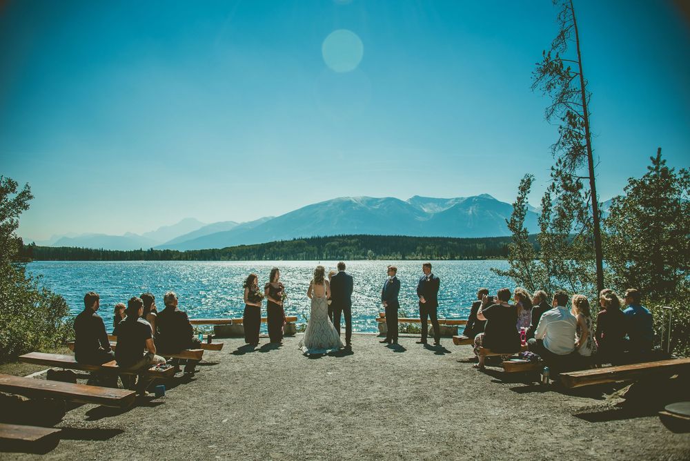 Outdoor micro wedding ceremony by a mountain lake with guests seated on benches, bridal party at the waterfront under a clear blue sky.