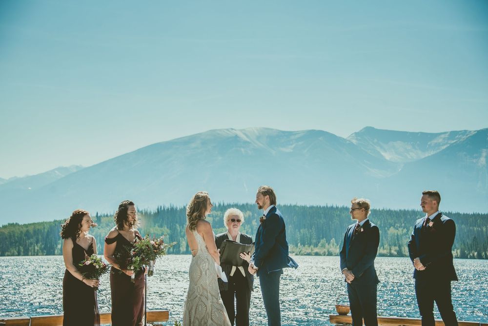 Micro Wedding ceremony on a lakeside dock with mountain backdrop, bridal party in black dresses and suits, officiant leading vows.
