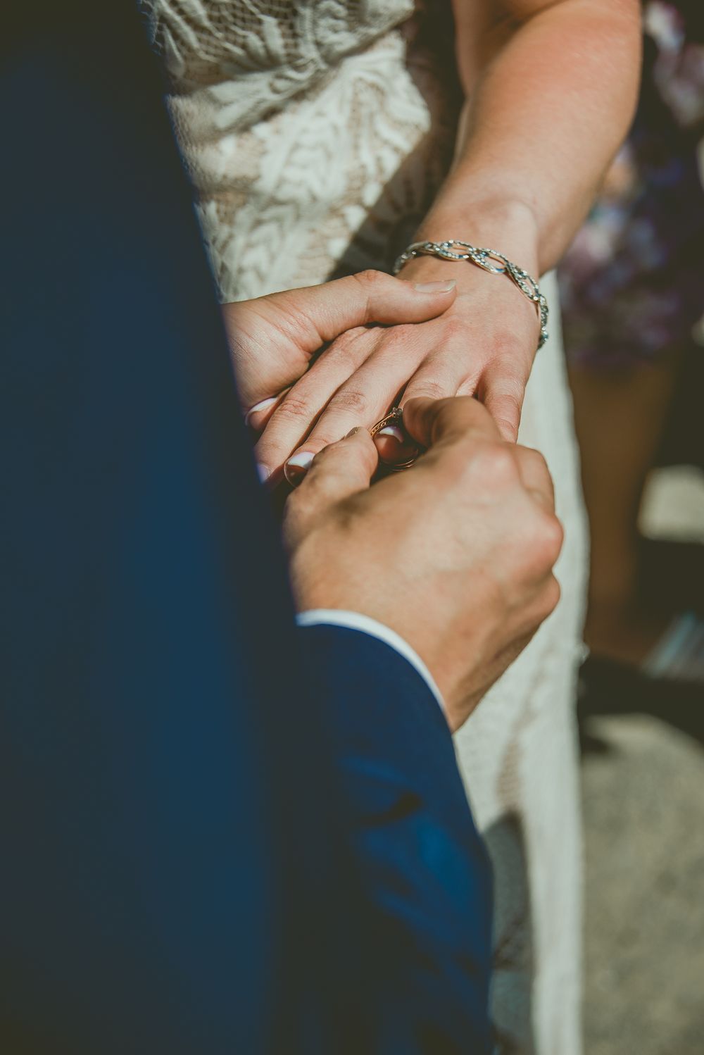Groom placing a wedding ring on brides finger during ceremony, bride wearing silver bracelet and lace dress.