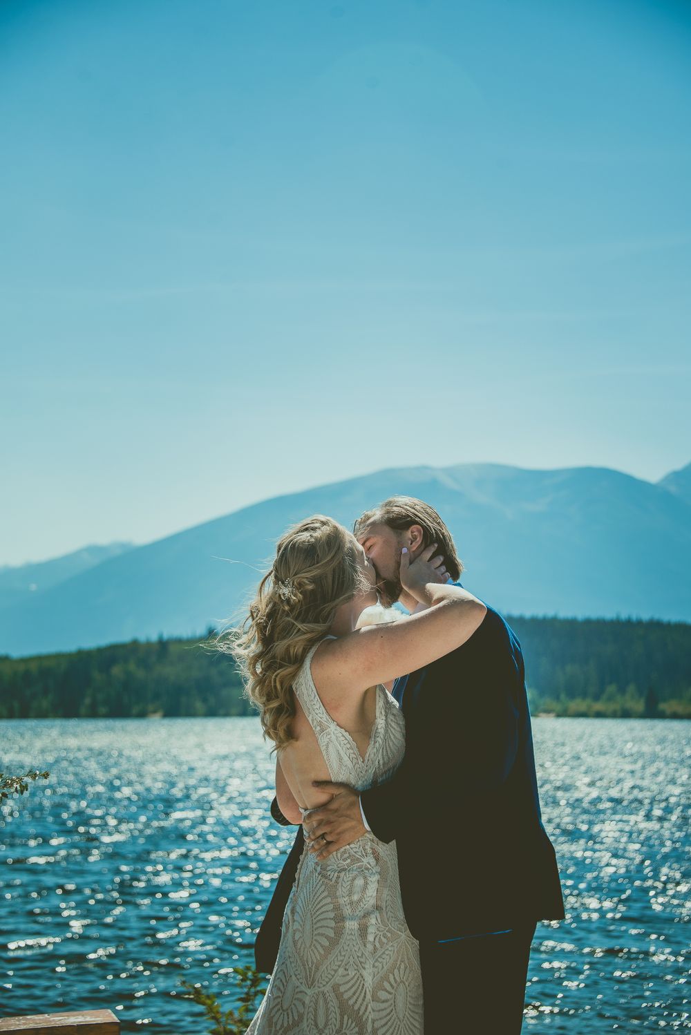 Bride and groom share first kiss at mountain lake wedding, bride in backless lace gown on sunny day.