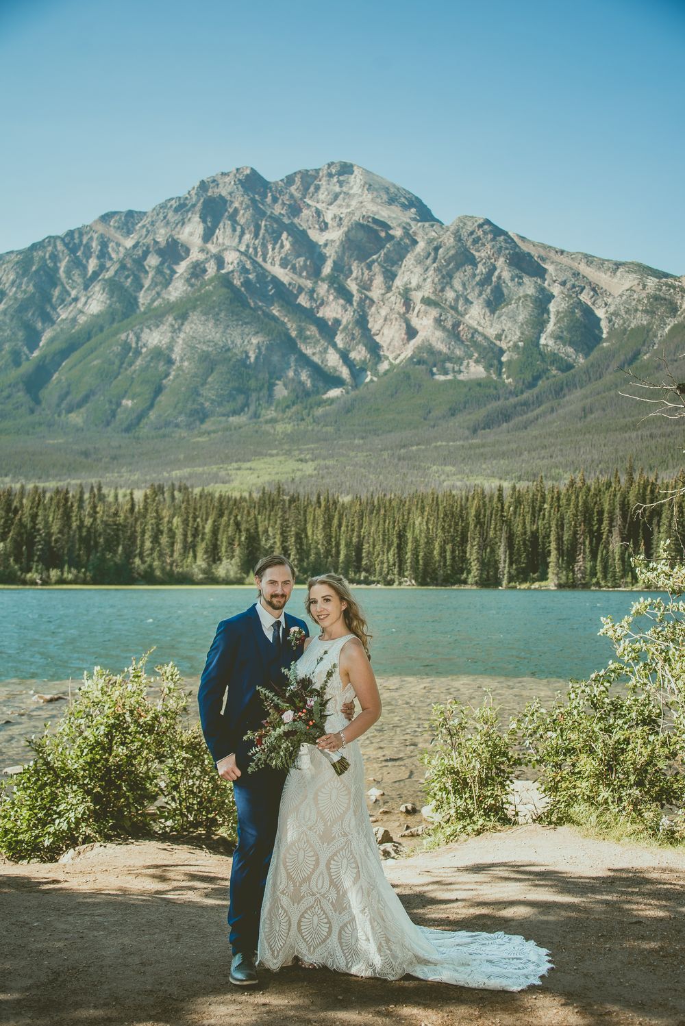 Bride and groom pose before turquoise mountain lake with Rocky Mountain backdrop.