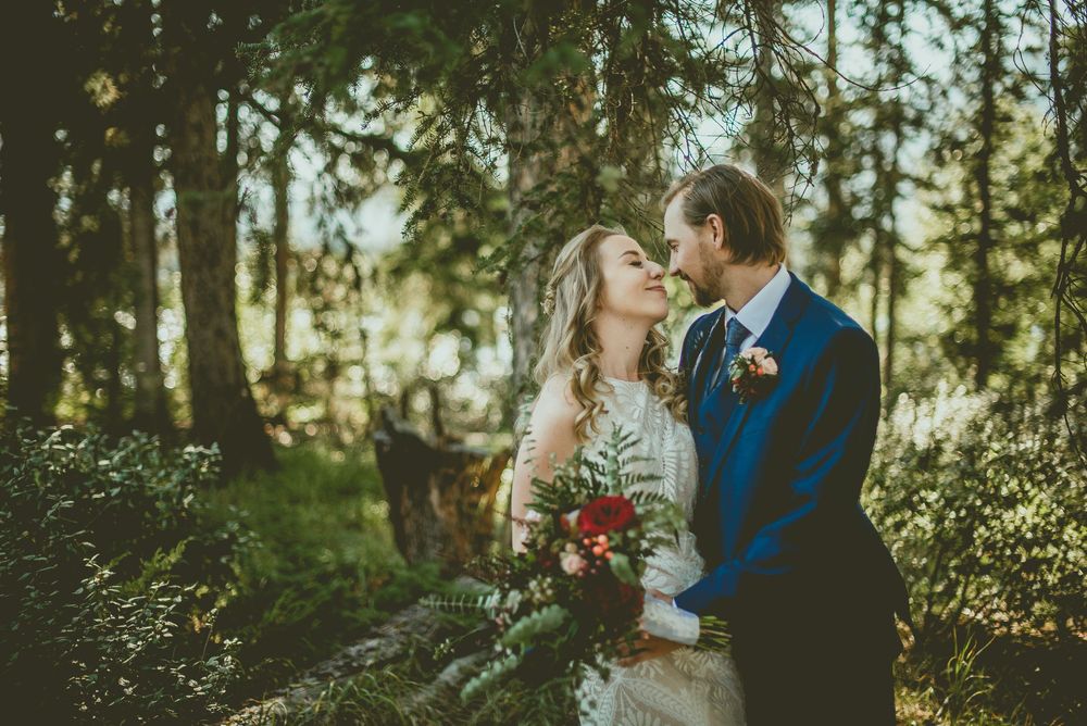 A bride and groom share a romantic moment in a sunlit forest, bride holding a red and green bouquet.