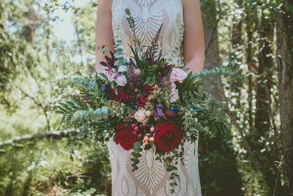 Bride holding a lush bohemian wedding bouquet with red peonies, ferns, and wildflowers in a forest setting.