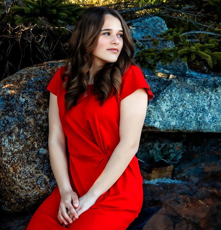 Young woman in red dress sitting on rocks near a waterfall in a serene outdoor portrait.