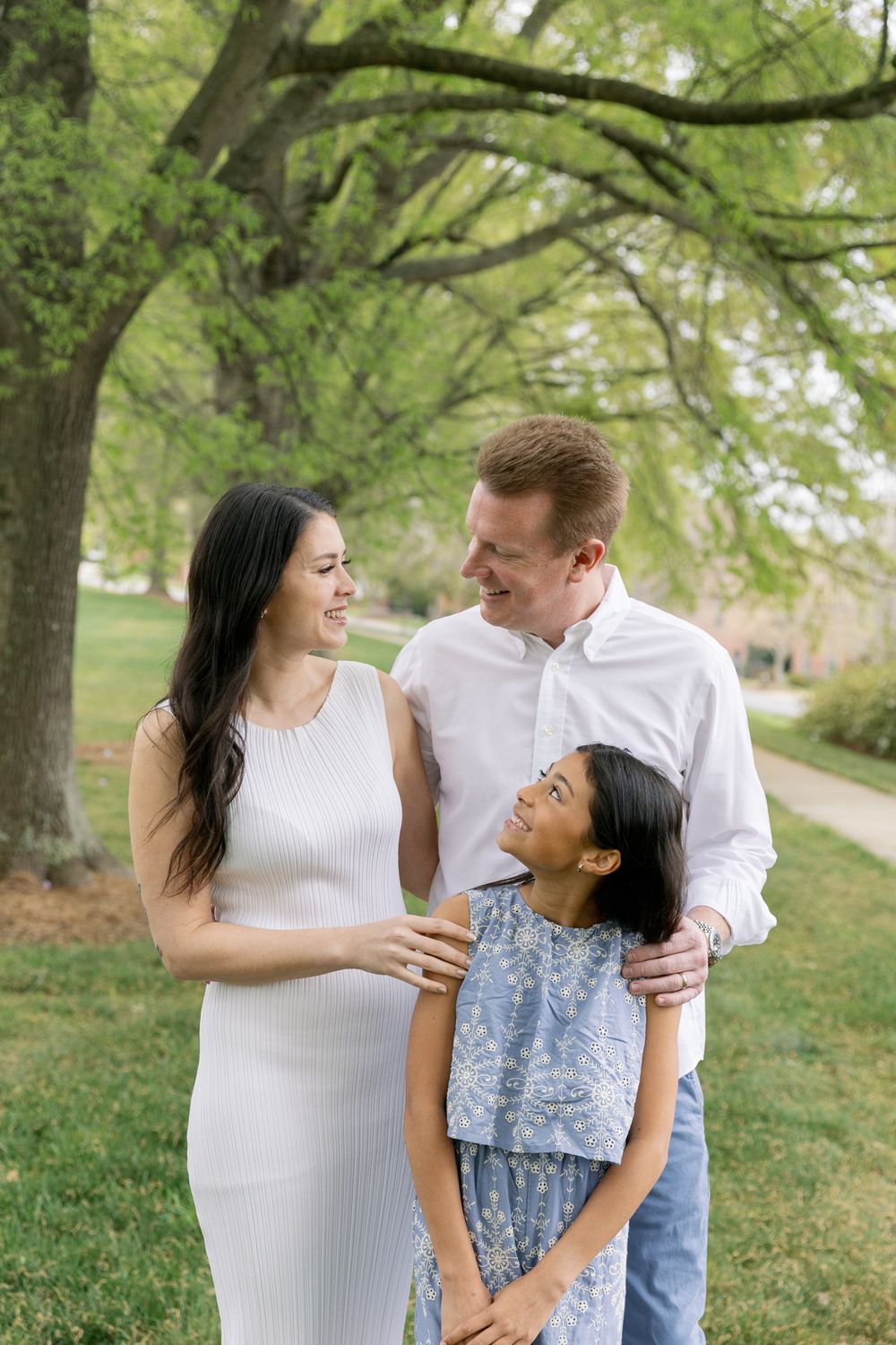 A happy family of three smiles together outdoors, with lush green trees in the background on a sunny day.