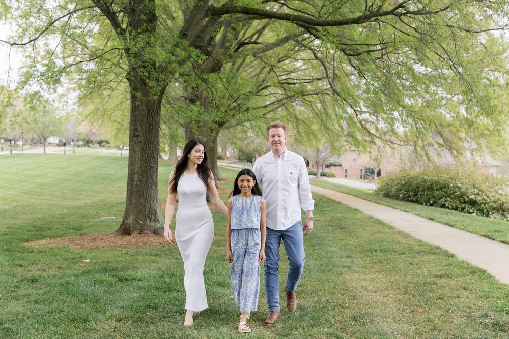 Happy family of three walking together on a green park path under large trees in spring.