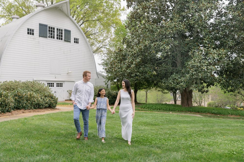 A happy family of three walks hand-in-hand on green grass near a white barn surrounded by lush trees.