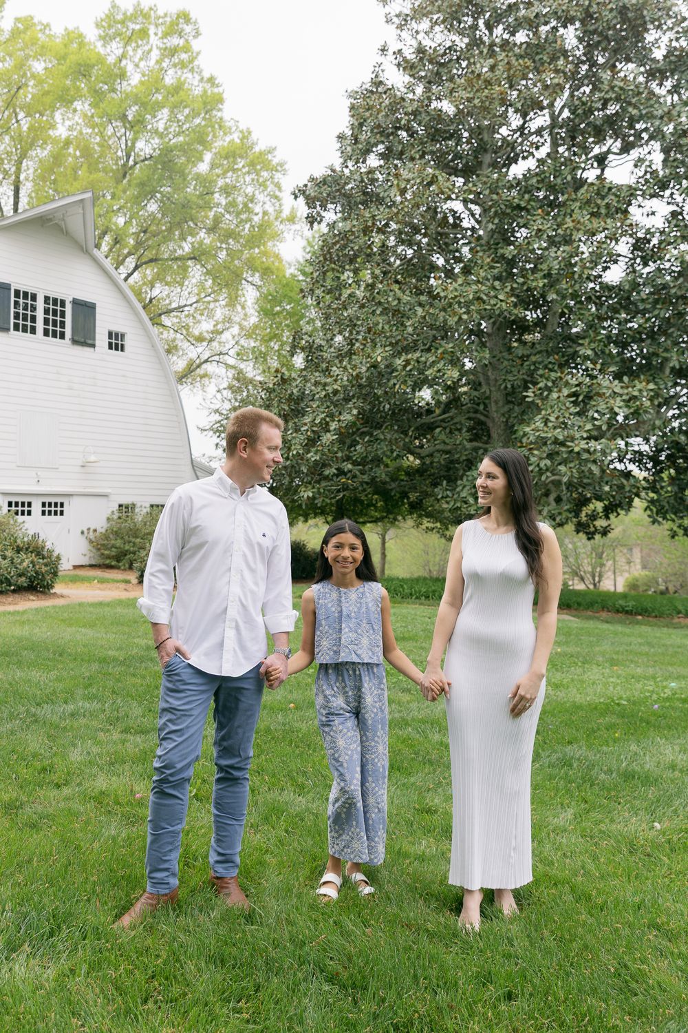 Happy family of three holding hands on green lawn beside white barn during spring outdoor photo session.