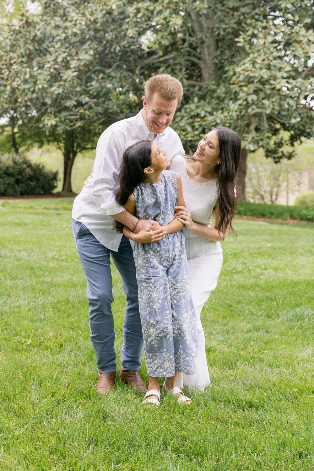 A happy family of three sharing a joyful moment outdoors on green grass with trees in the background.