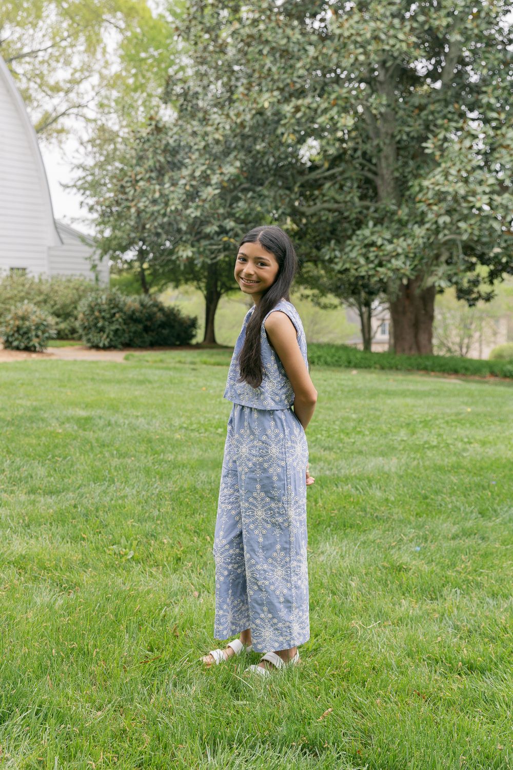 A young girl in a blue floral jumpsuit smiles over her shoulder while standing on a green lawn.