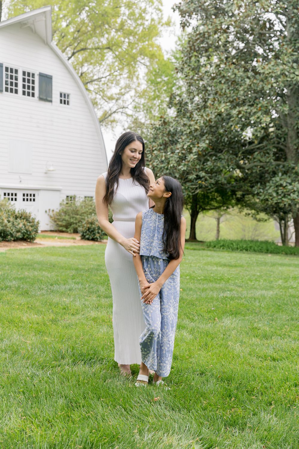 A mother and daughter share a tender moment standing together on a lush green lawn near a white barn.