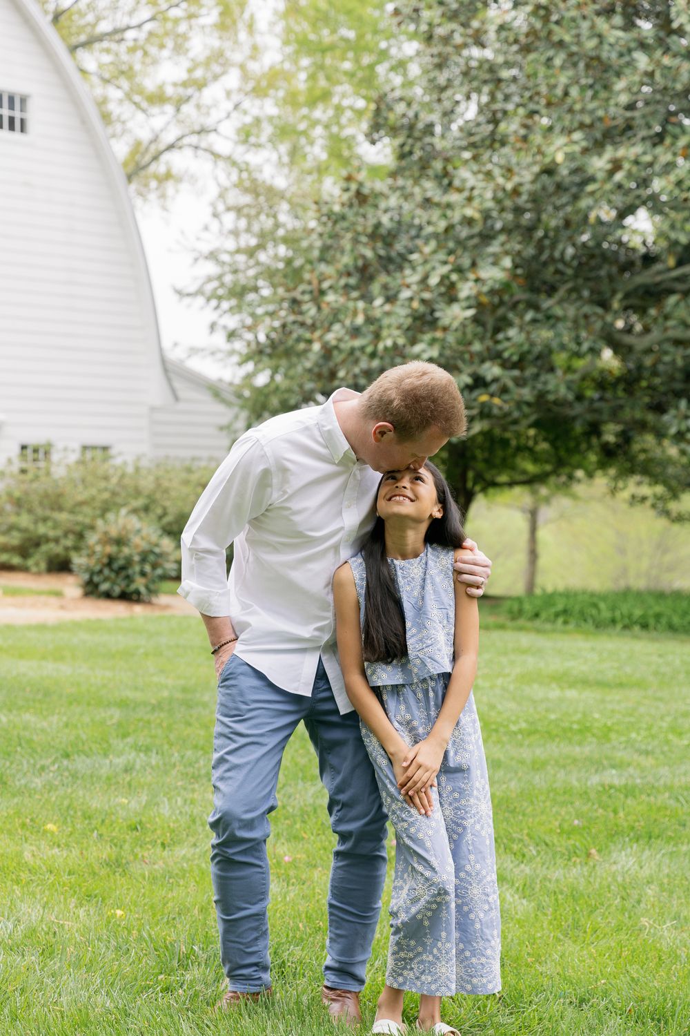 Tall man tenderly kissing young girl on forehead while standing on green lawn near white barn.
