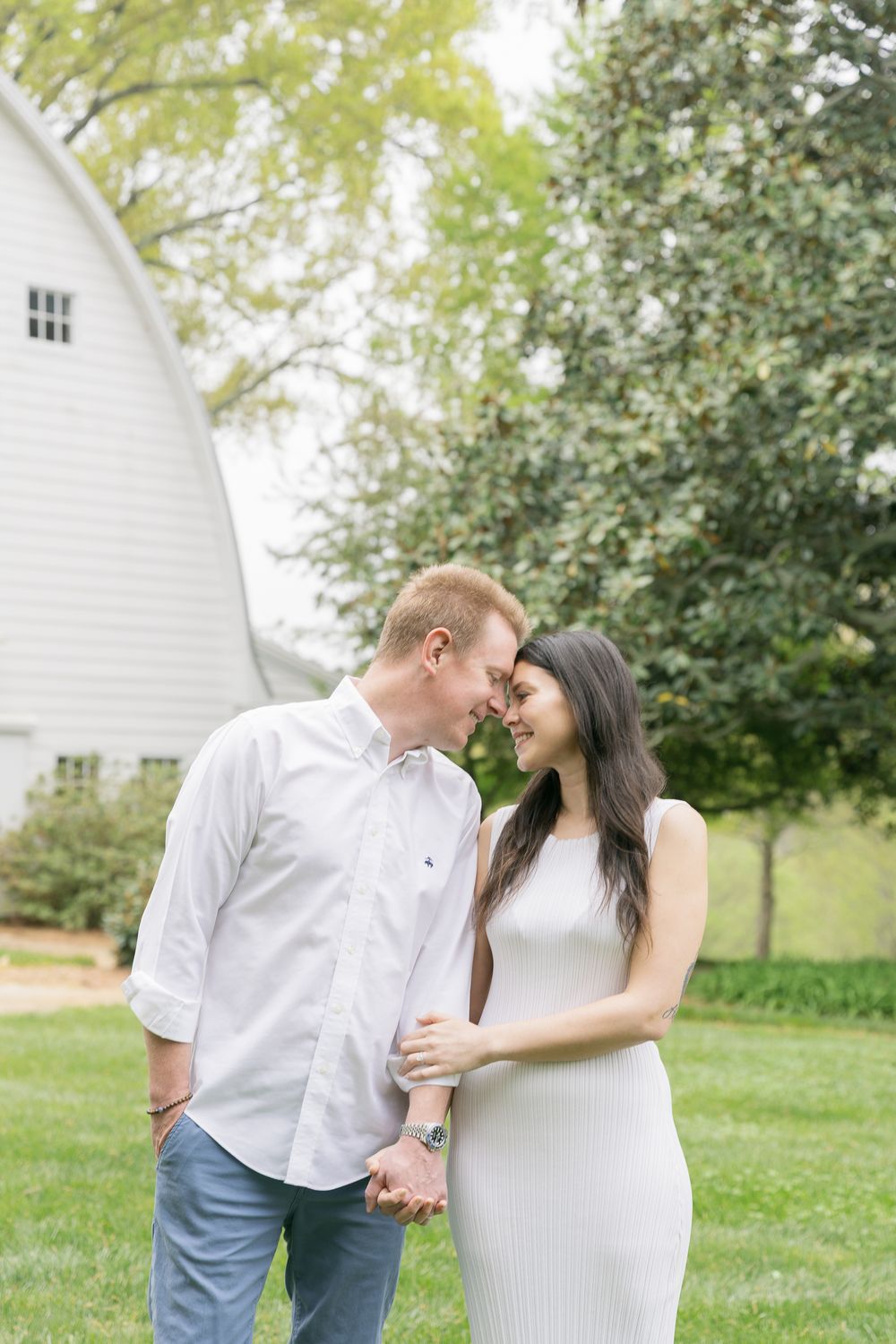 A couple shares a romantic forehead touch while holding hands outdoors near a white barn surrounded by lush greenery.