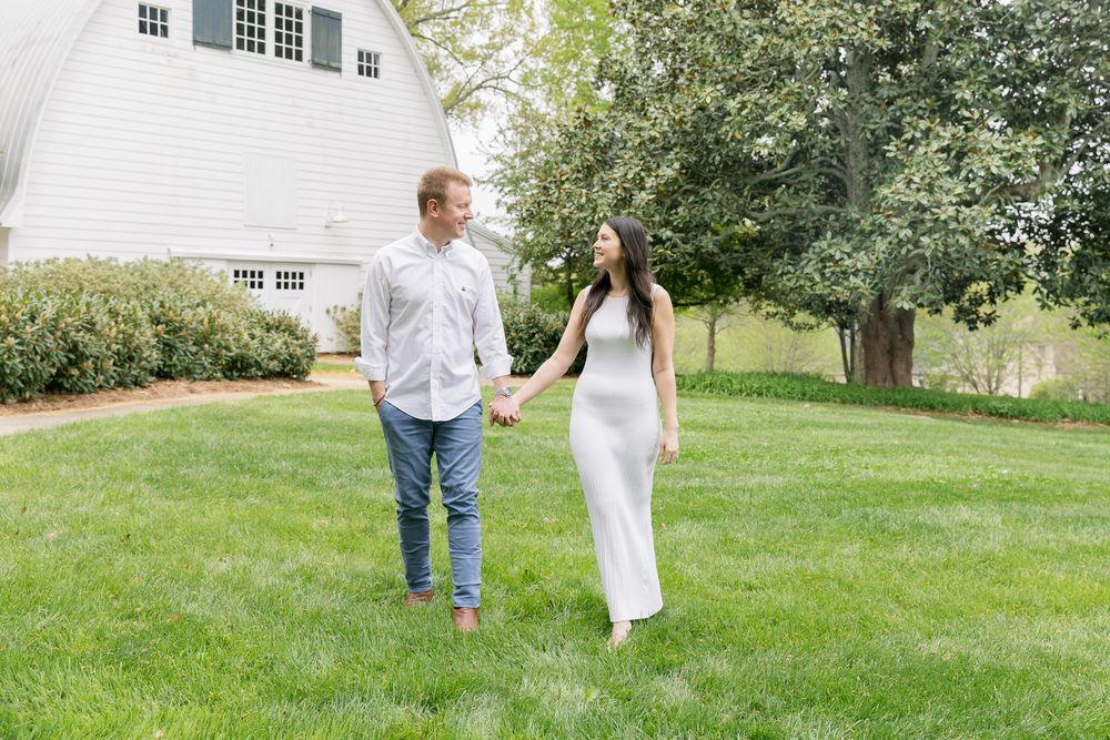 A couple holds hands while walking across a green lawn beside a white barn on a sunny day.