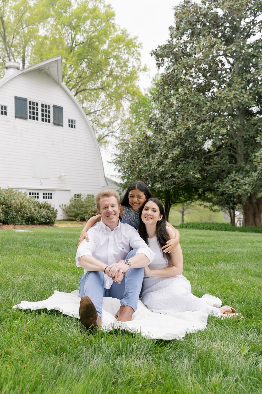 Happy family of three sitting on white blanket in green yard near white barn on spring day.