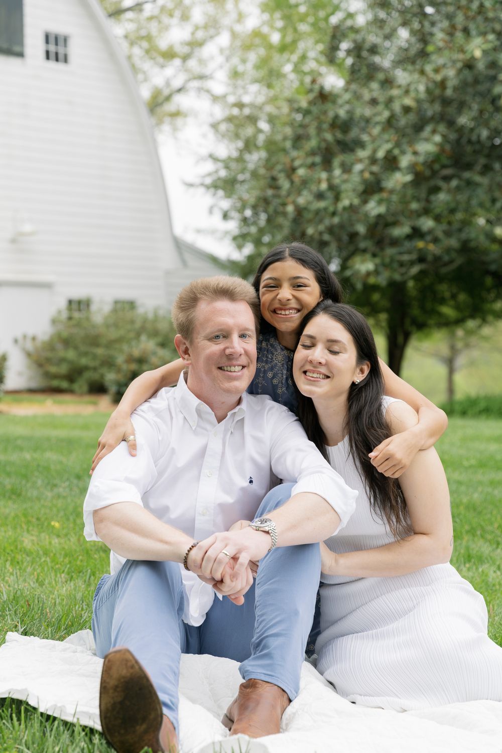 Joyful family of three sitting together on white blanket on grass in front of white barn.