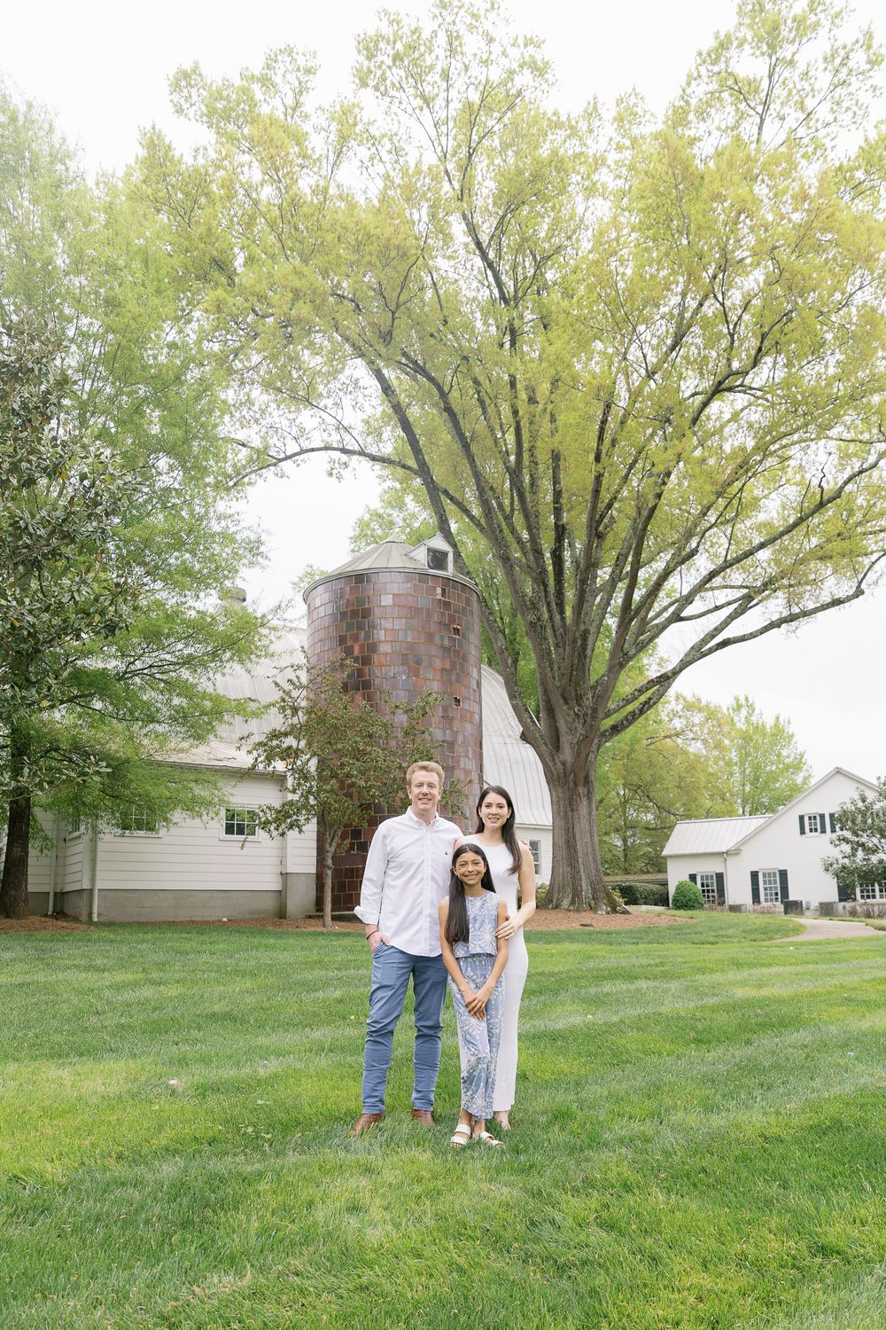 A family of three poses together on a green lawn with a rustic silo and large tree in the background.