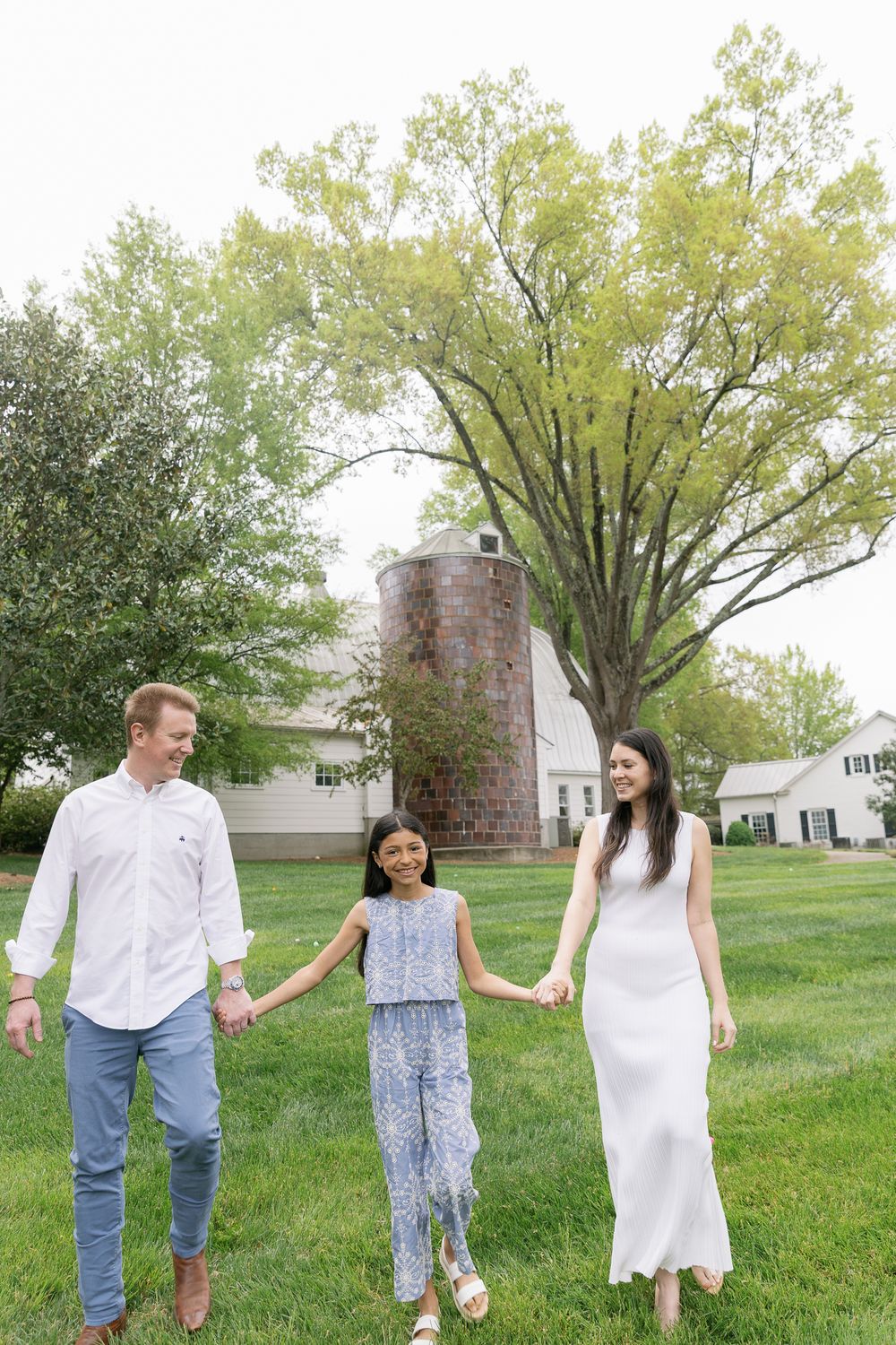Family of three holding hands walking on green lawn near barn and silo.