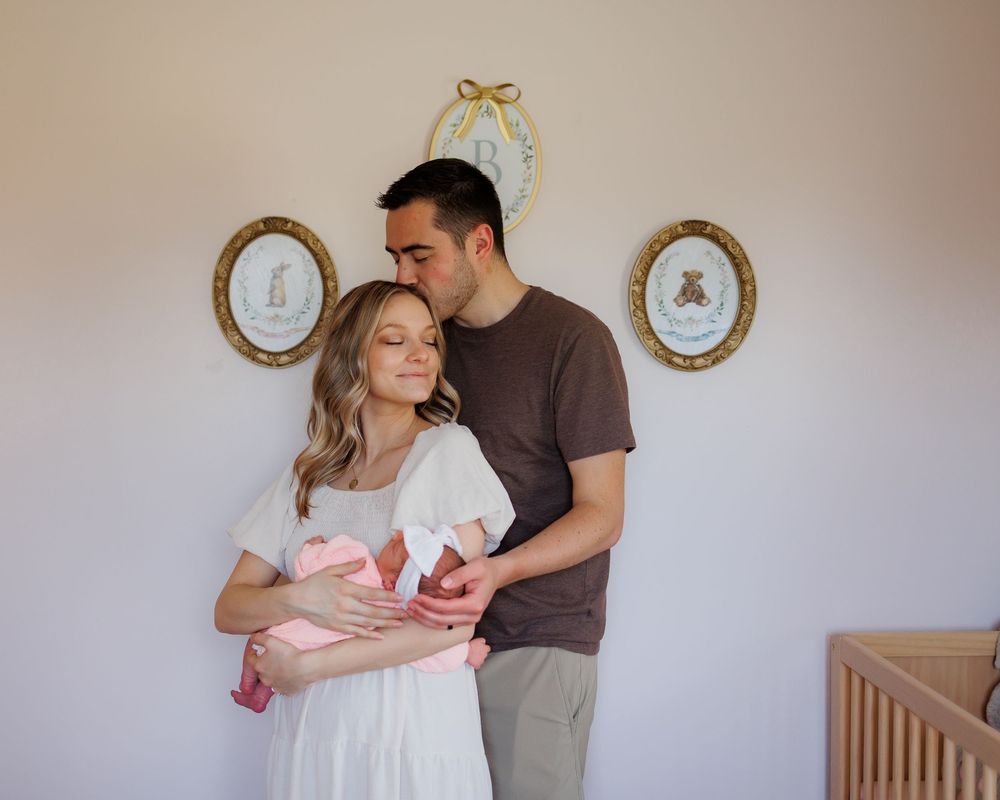 New parents couple holding newborn baby in pink blanket in a nursery room with decorative wall frames.
